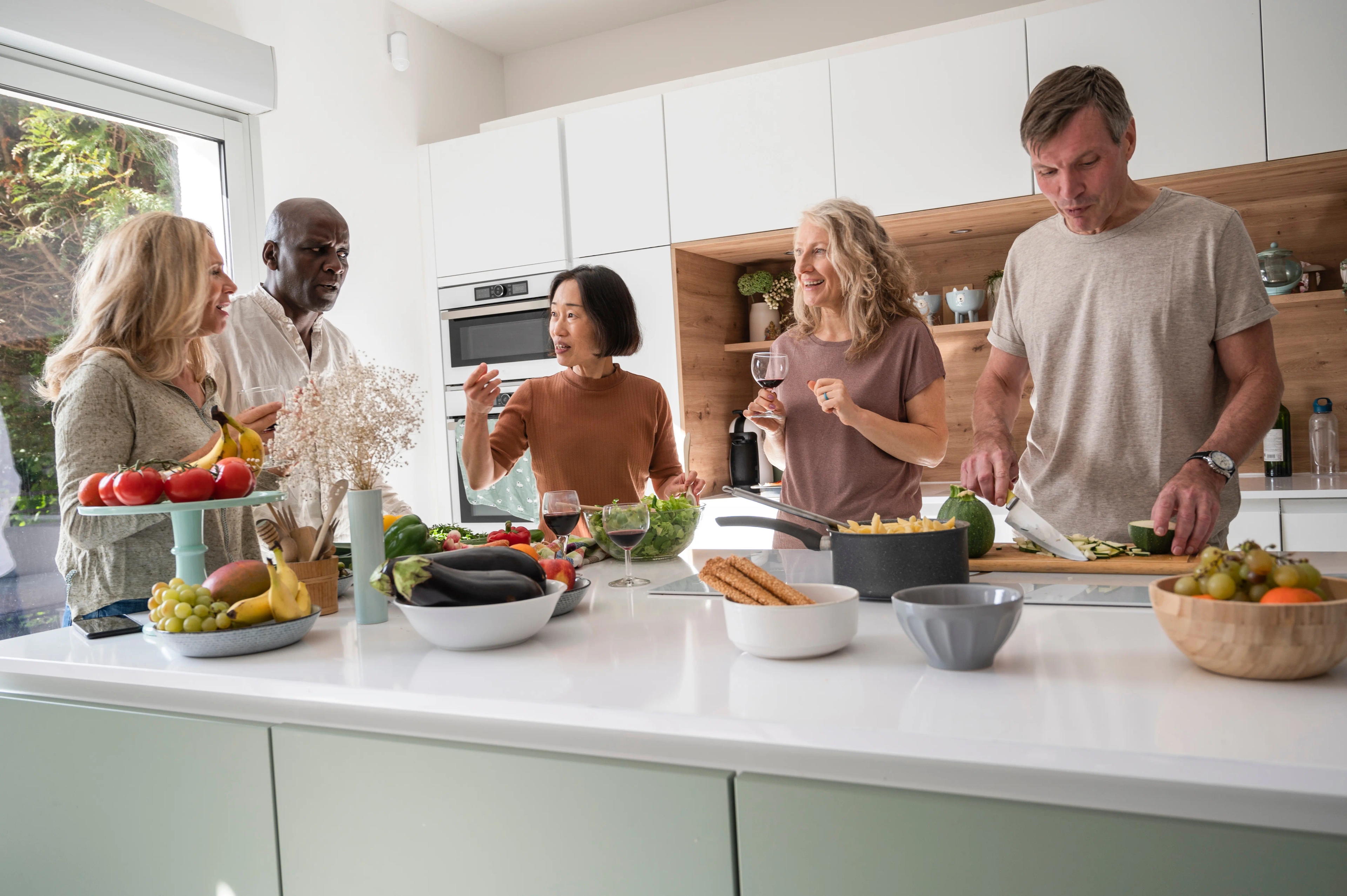 Five adults chatting and preparing a meal together in a bright, modern kitchen with fresh produce on the countertop